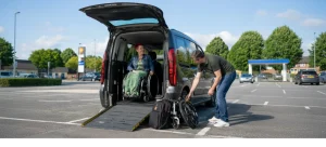 A female wheelchair user sat inside a wheelchair accessible vehicle with the ramp deployed. Husband is sorting luggage at the side of the van.